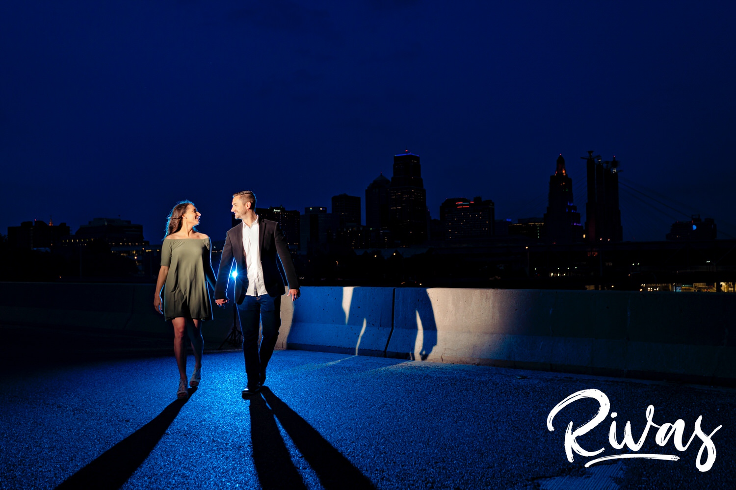 A colorful photo taken just after dark on the Summit Street Bridge looking back at the Kansas City skyline of an engaged couple walking towards the camera as they hold hands during their summer engagement session. 