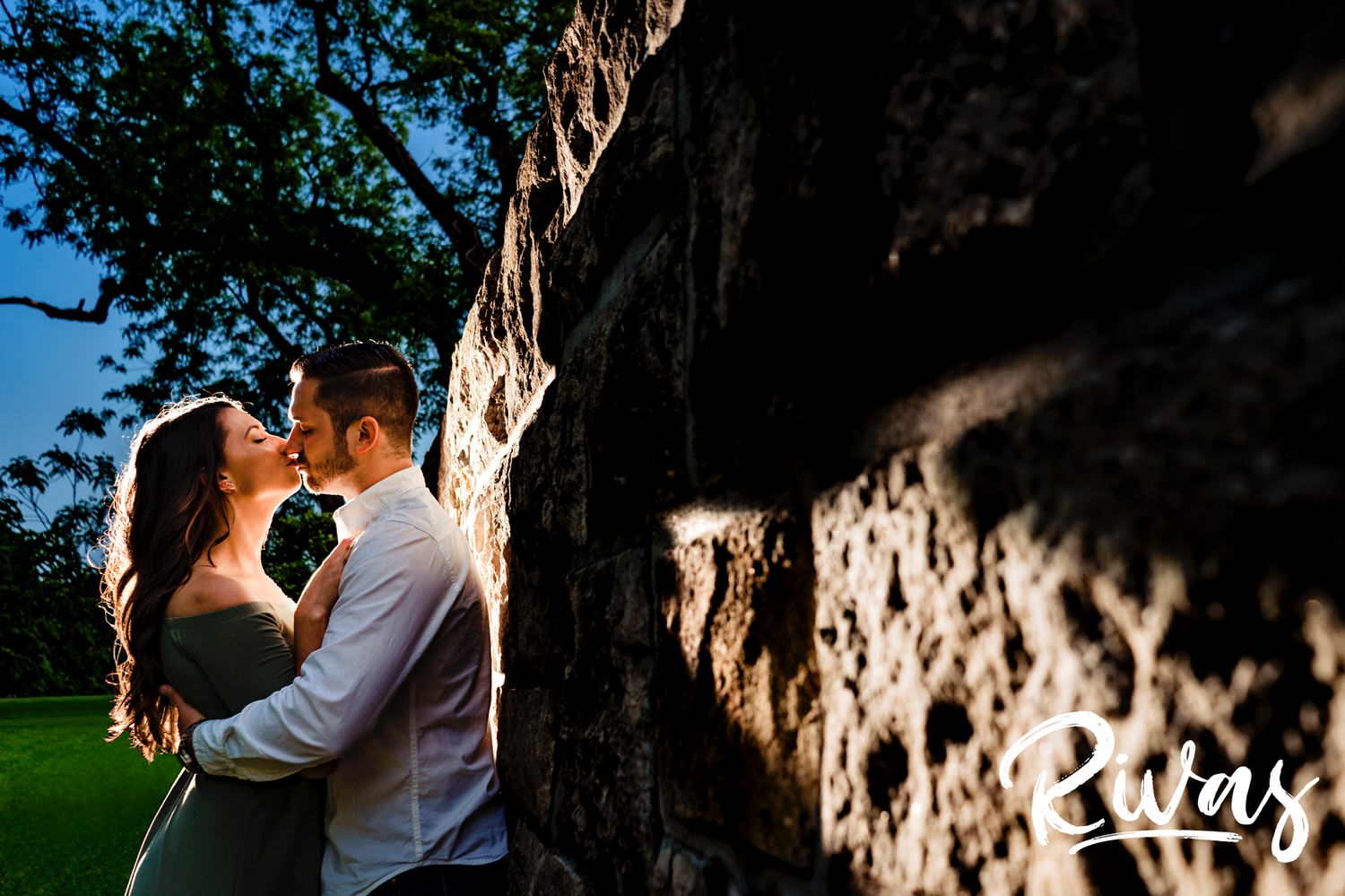 A colorful portrait of an engaged couple leaning up against a stone wall sharing a kiss during their engagement session at Loose Park in Kansas City. 