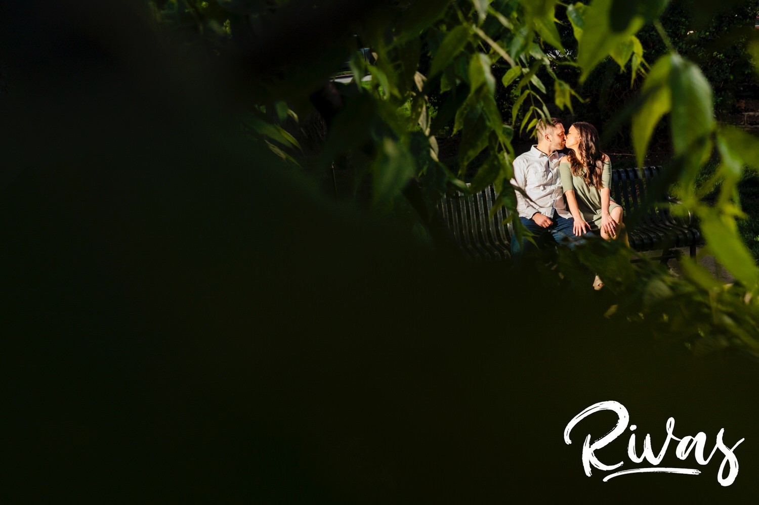 A colorful picture taken through the trees of an engaged couple sitting on a park bench during their summer engagement session at Loose Park in Kansas City. 
