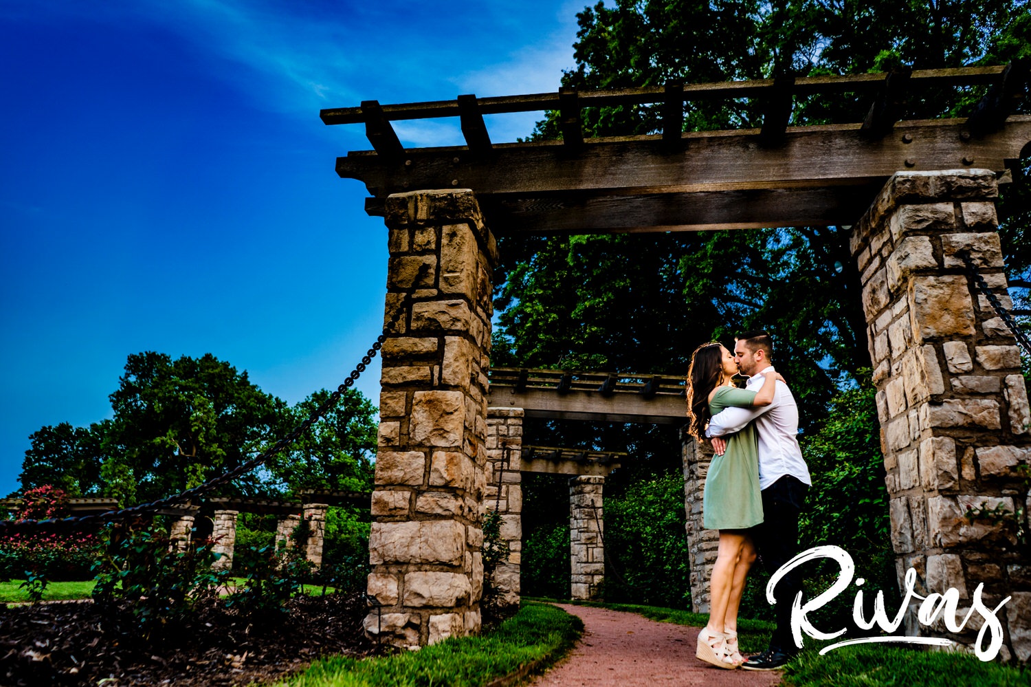A colorful portrait of an engaged couple sharing and embrace and kissing underneath the stone arbor in Kansas City's Loose Park Rose Garden. 