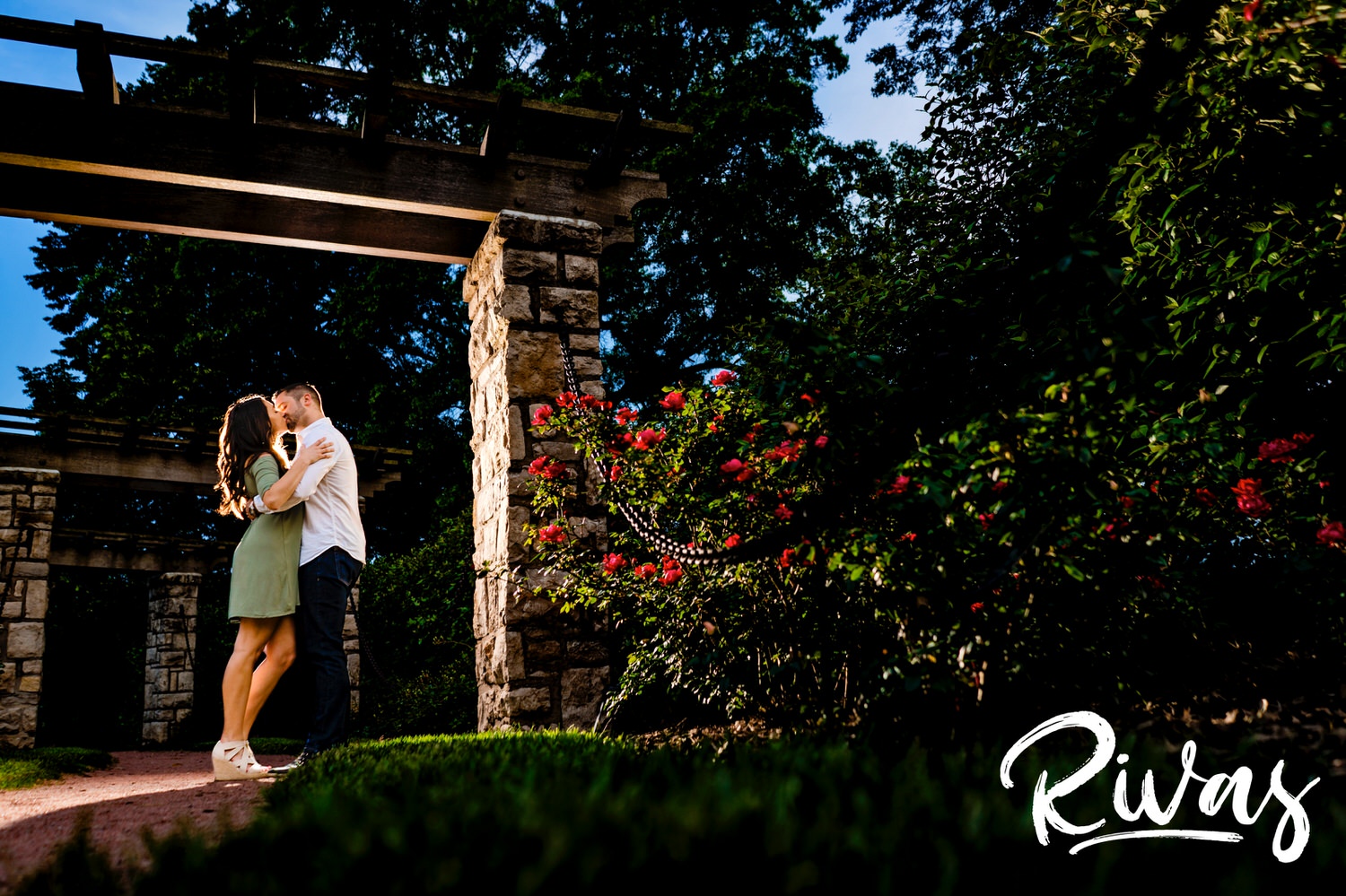A colorful portrait of an engaged couple sharing and embrace and kissing underneath the stone arbor in Kansas City's Loose Park Rose Garden.