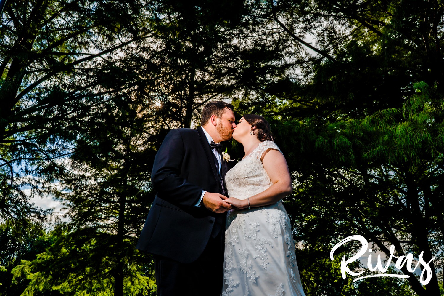 A colorful picture of a bride and groom standing together, holding hands and sharing a kiss underneath a canopy of green trees on their summer wedding day in Lawrence, Kansas. 