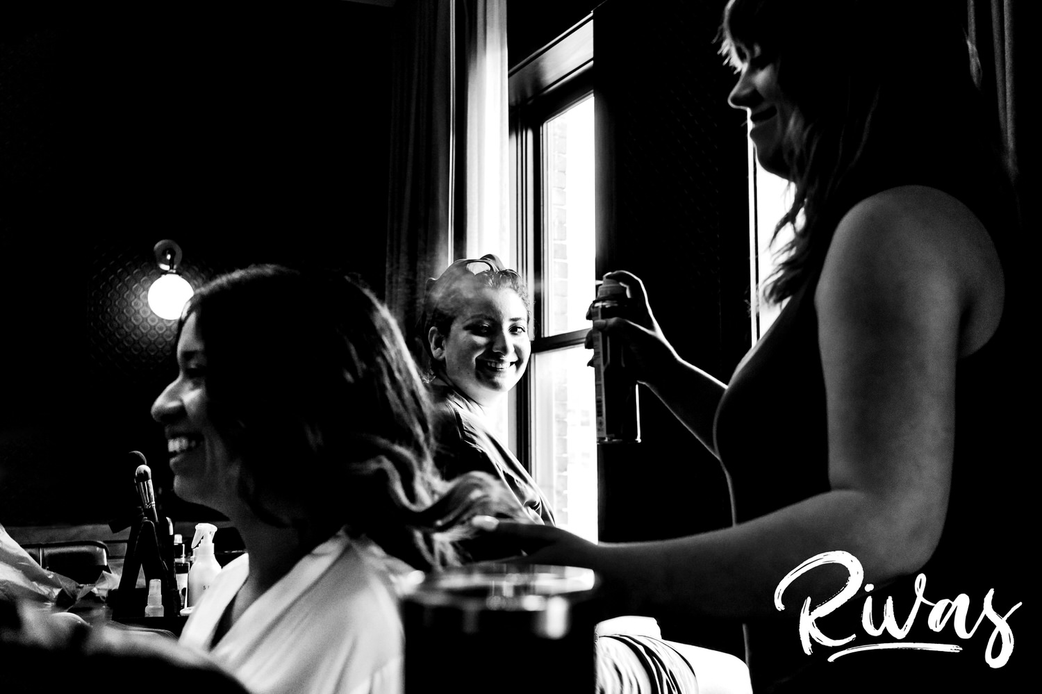 A candid black and white picture of a bride's sister, smiling at her over her shoulder as they both get hair and make-up done on the morning of a KC wedding. 