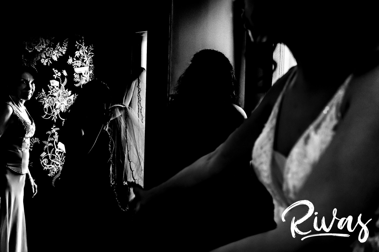 A dramatic black and white picture of a bride reaching out for her veil as her mom smiles around the corner in the background on the morning of a Kansas City wedding. 