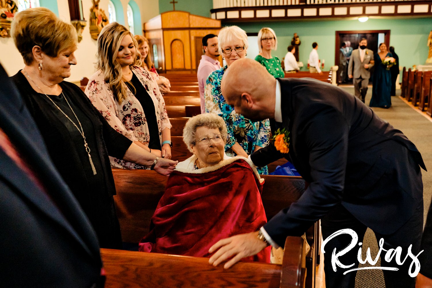 A candid picture of a groom leaning in to kiss his grandma just after she's been seated for his wedding ceremony. 