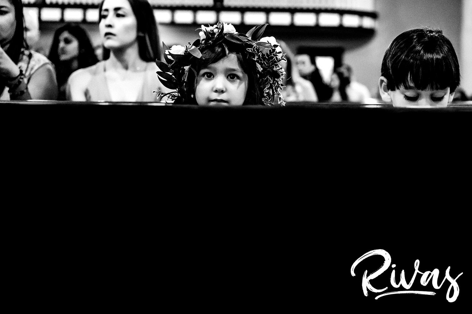 A candid black and white picture of a flower girl in a flower crown peeking over the back of a pew during a wedding ceremony in Kansas City. 