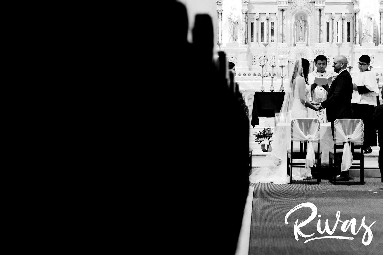 A black and white picture taken from the back of the church of a bride and groom standing at the front of the church during their wedding ceremony. 