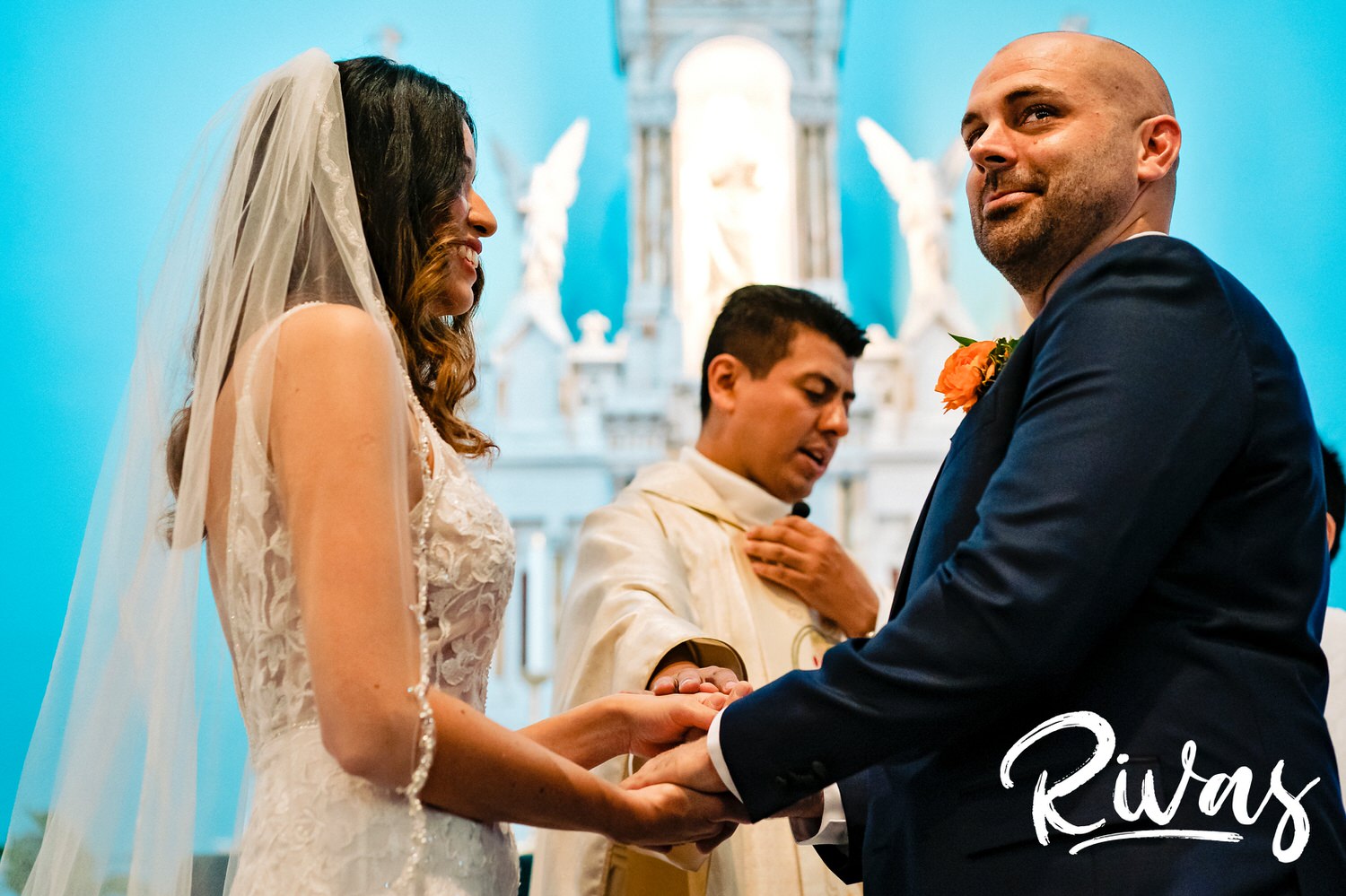 A candid picture taken during a wedding ceremony of a groom holding his bride's hand and looking out at their guests and smiling during their wedding ceremony in Kansas City. 