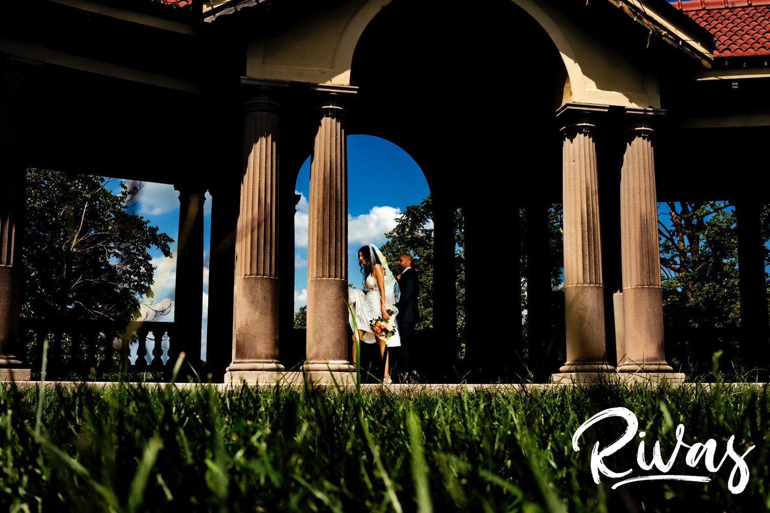 A candid, wide picture of a bride and groom walking underneath a set of columns against a bright blue sky on the afternoon of their wedding in Kansas City. 