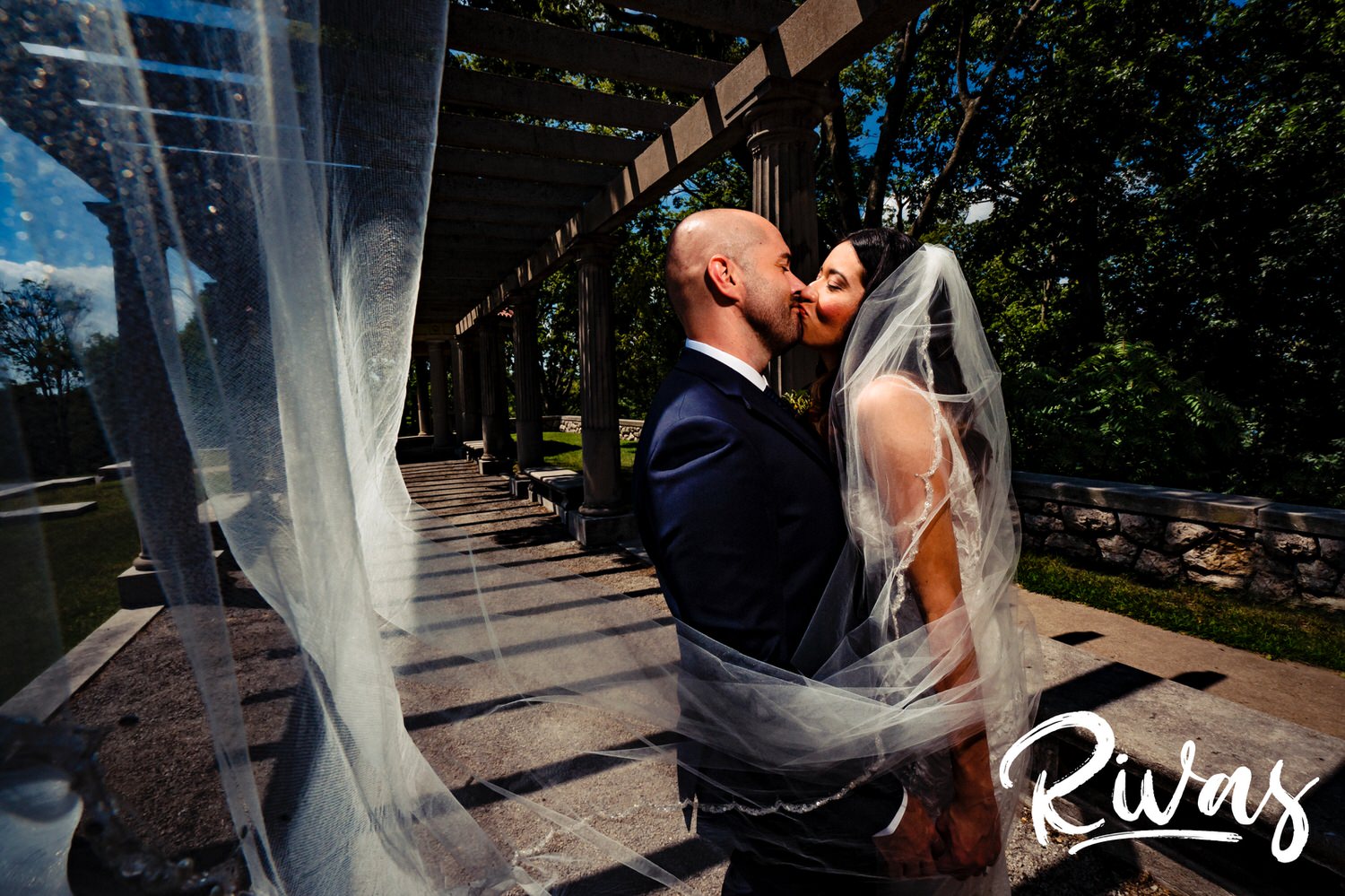 A candid portrait of a bride and groom sharing a kiss as the bride's veil blows in the wind on the afternoon of their Kansas City wedding. 