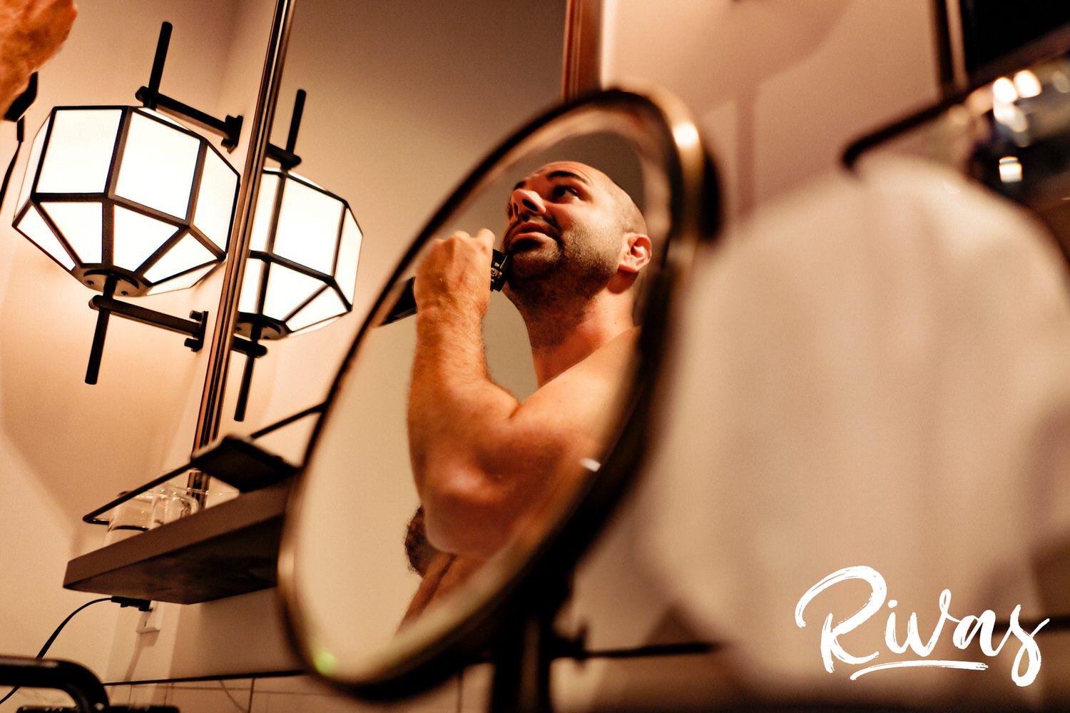 A candid picture of a groom shaving on the morning of his wedding, visible in a round mirror in a hotel bathroom. 