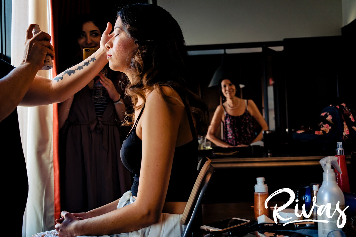 A candid picture of a bride getting her hair and make-up done with her mom in the background smiling on the morning of her wedding in Kansas City. 