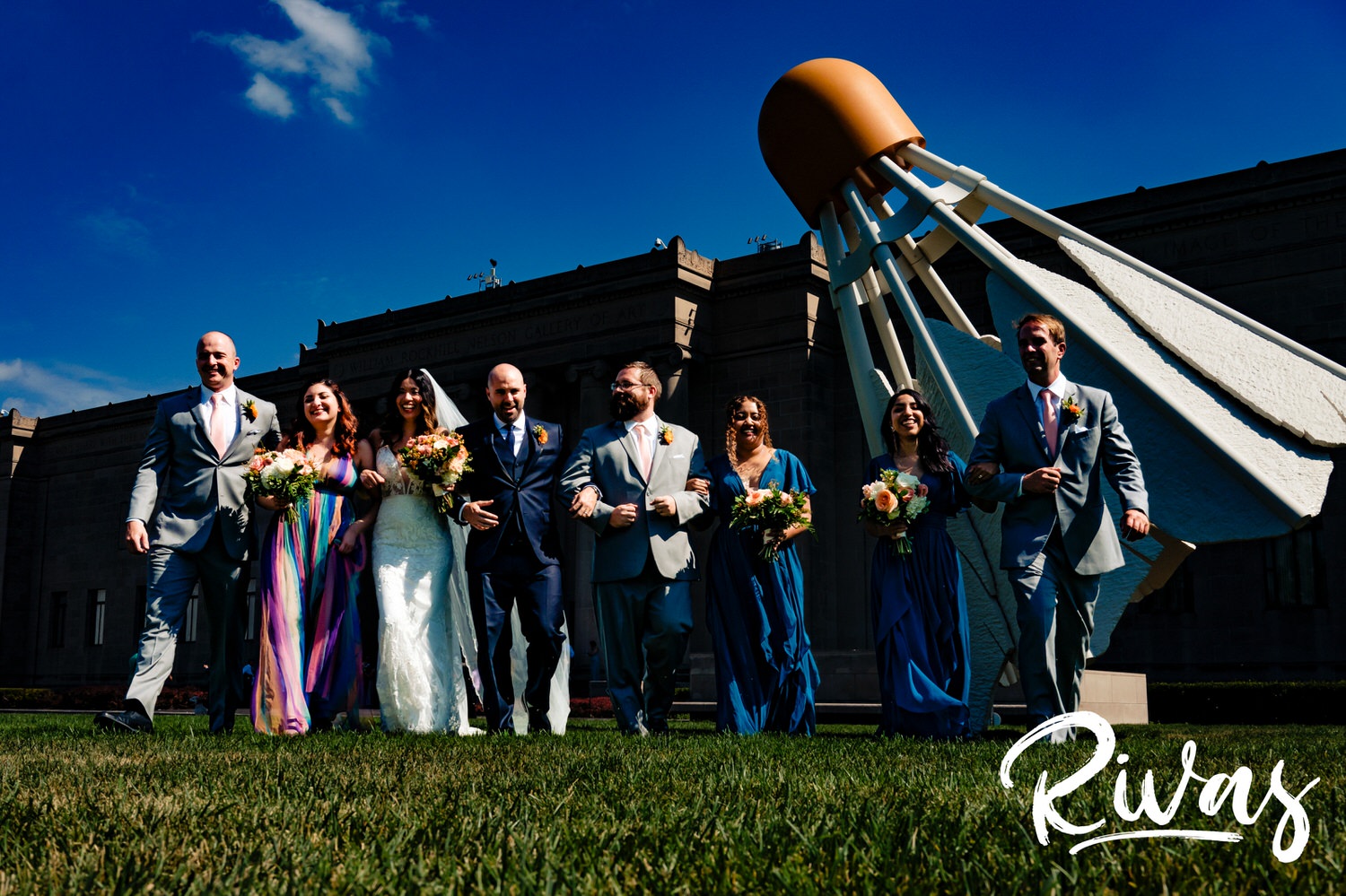 A bright, candid picture of a wedding party walking arm in arm across the green lawn of The Nelson Atkins Museum of Art with a Shuttlecock visible in the background. 