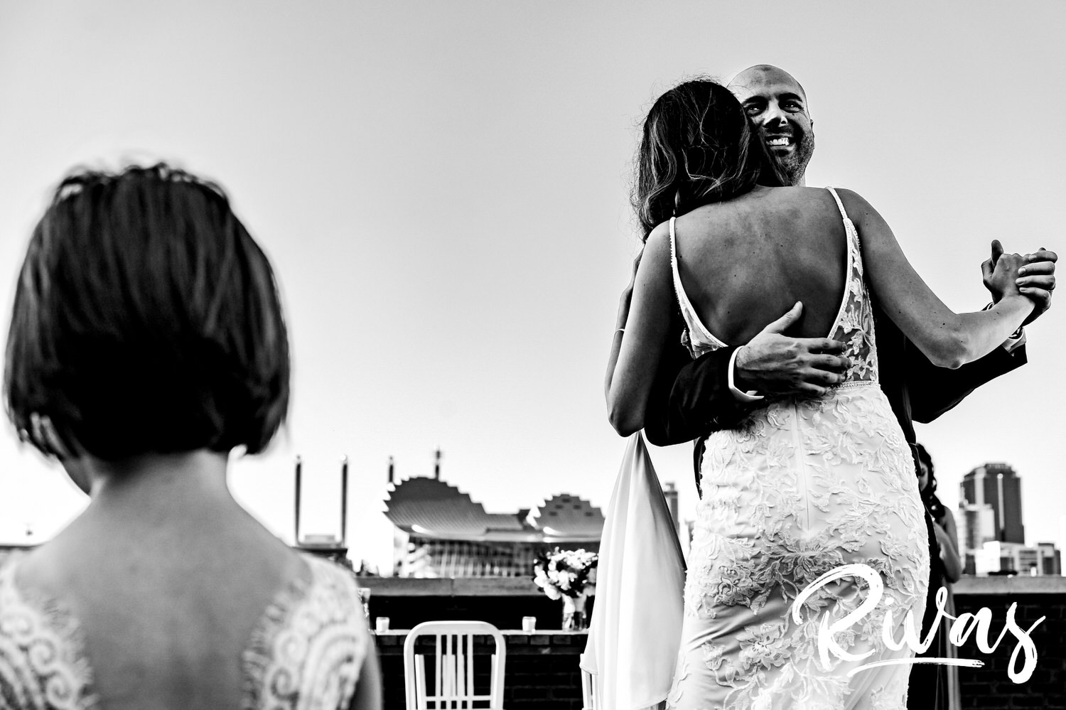 A candid black and white picture of a bride and groom sharing their first dance in front of the Kansas City skyline as a flower girl watches on. 