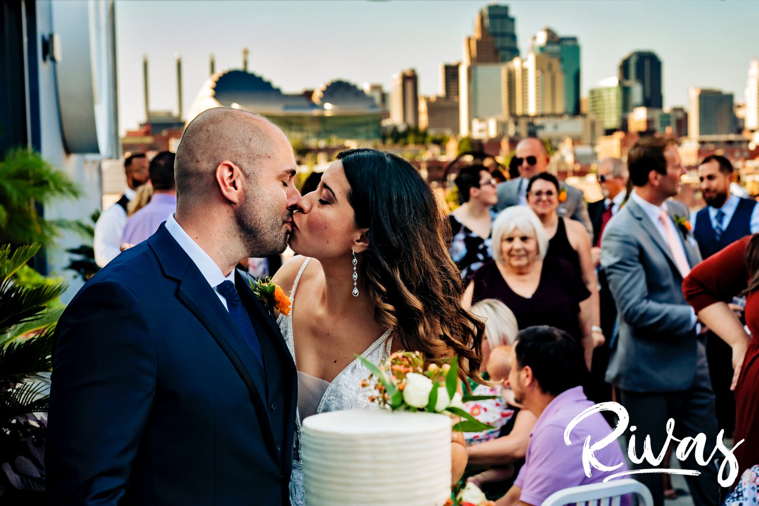 A colorful, candid picture of a bride and groom sharing a kiss as they cut their wedding cake in front of the Kansas City Skyline on their wedding day.