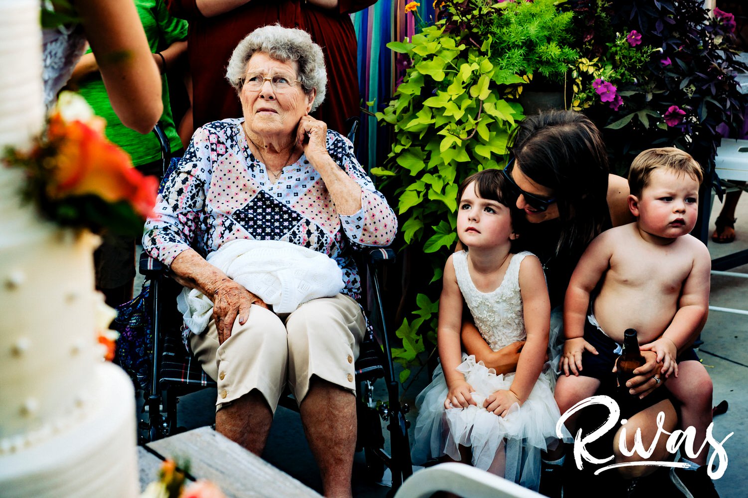 A colorful, candid picture of a groom's grandma, a flower girl, and a ring bearer with no shirt on watching a bride and groom cut the cake during their wedding reception in Kansas City. 