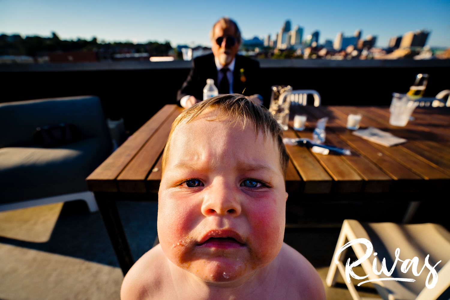 A very candid, very close-up picture of a tiny ring bearer, bright red from the sun, staring directly into the camera lens as the father of the groom eats a piece of cake in the background in front of the Kansas City skyline. 
