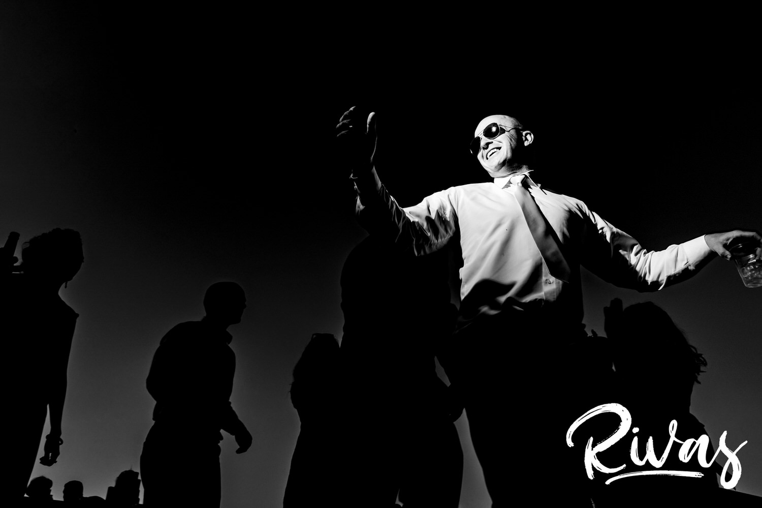 A candid black and white picture of a bald man in sunglasses dancing during a wedding reception, with the silhouettes of fellow dancers visible behind him during a wedding reception in Kansas City. 