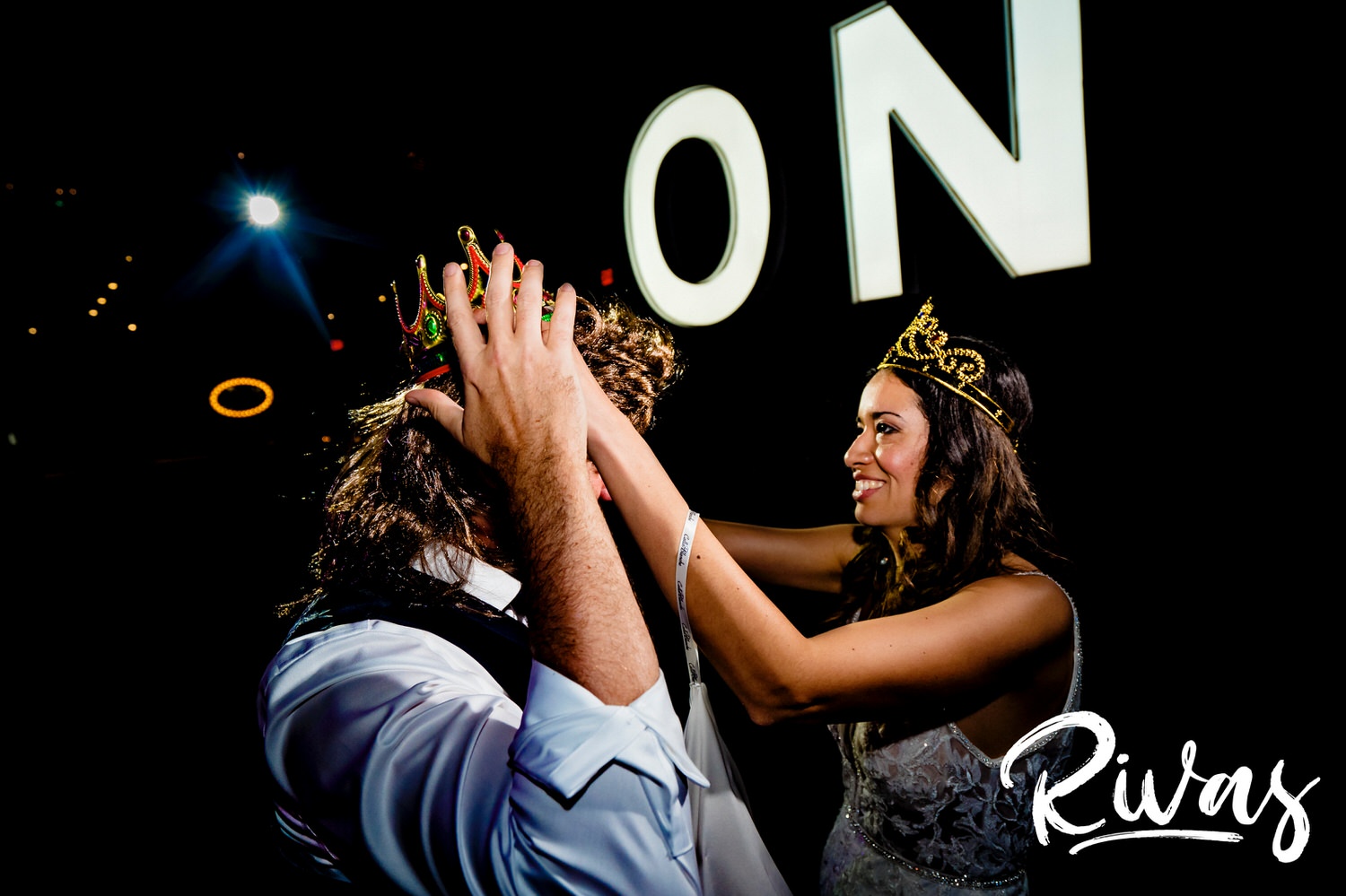 A candid picture of a bride in a crown adjusting a crown on her groom's head, who is wearing a mullet wig, during their wedding reception in Kansas City. 