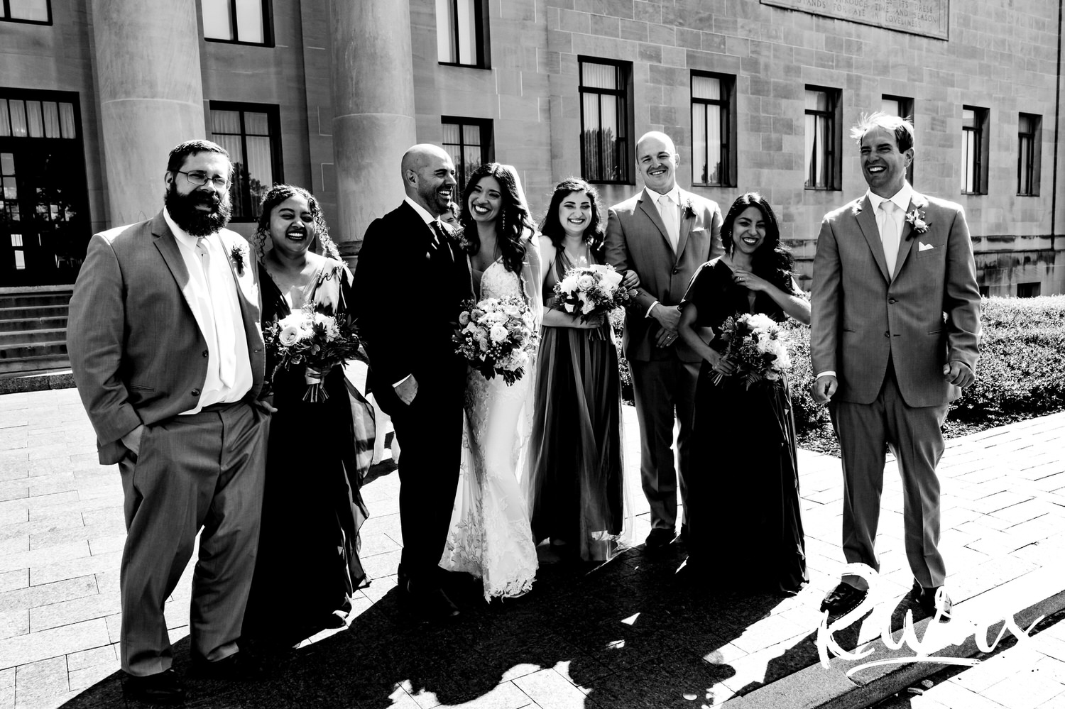 A candid black and white picture of a wedding party walking arm in arm across the front patio of The Nelson Atkins Museum of Art on their Kansas City wedding day. 