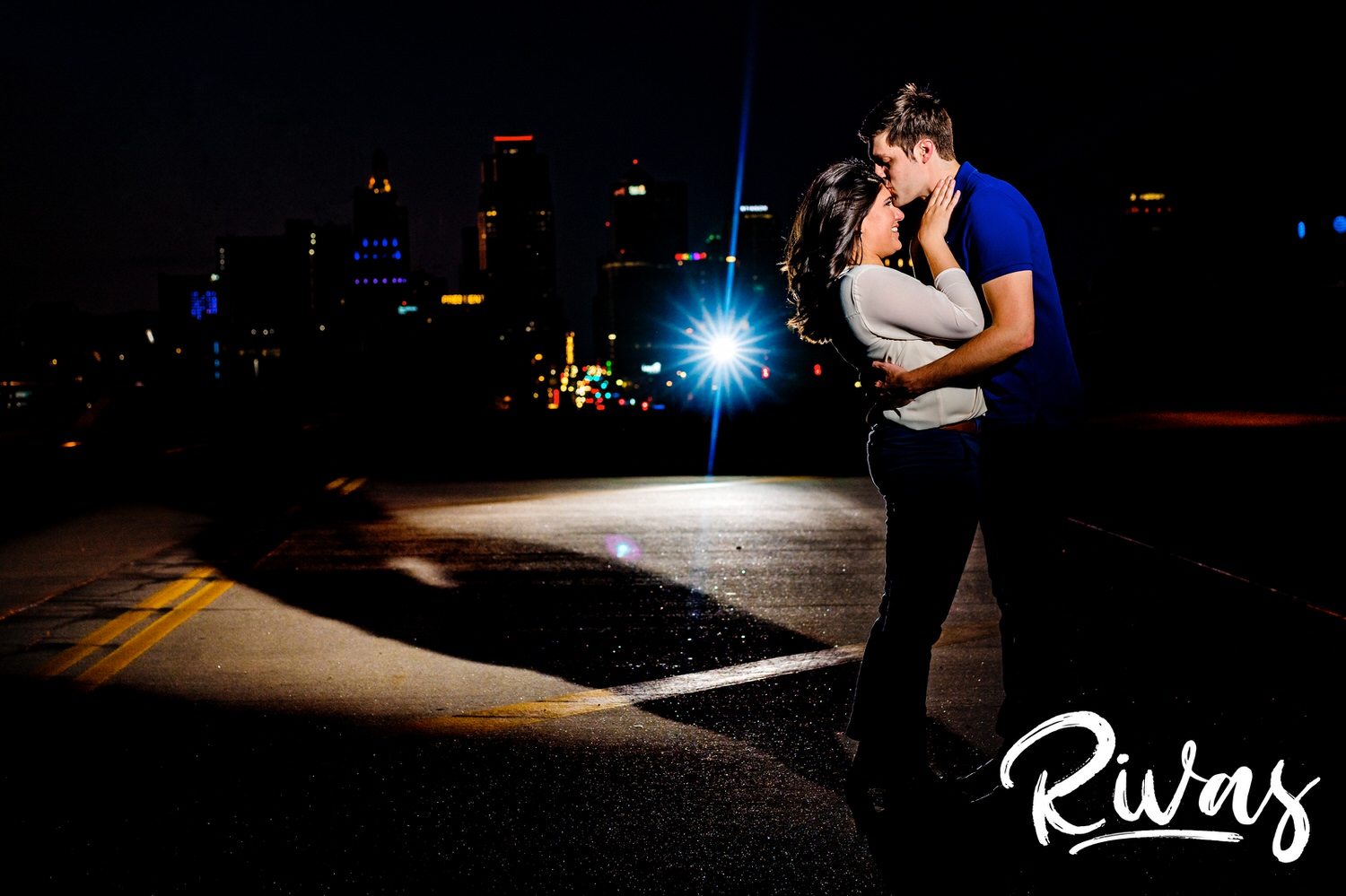 An vibrant, bold picture of an engaged couple standing in the middle of Main Street in Kansas City during their nighttime engagement session, sharing an embrace and kiss with the KC skyline visible in the background. 