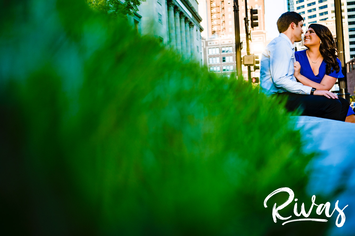 A bright, candid picture of an engaged couple holding hands, sitting on a ledge in front of a bright green lawn during their fall engagement session in downtown Kansas City. 
