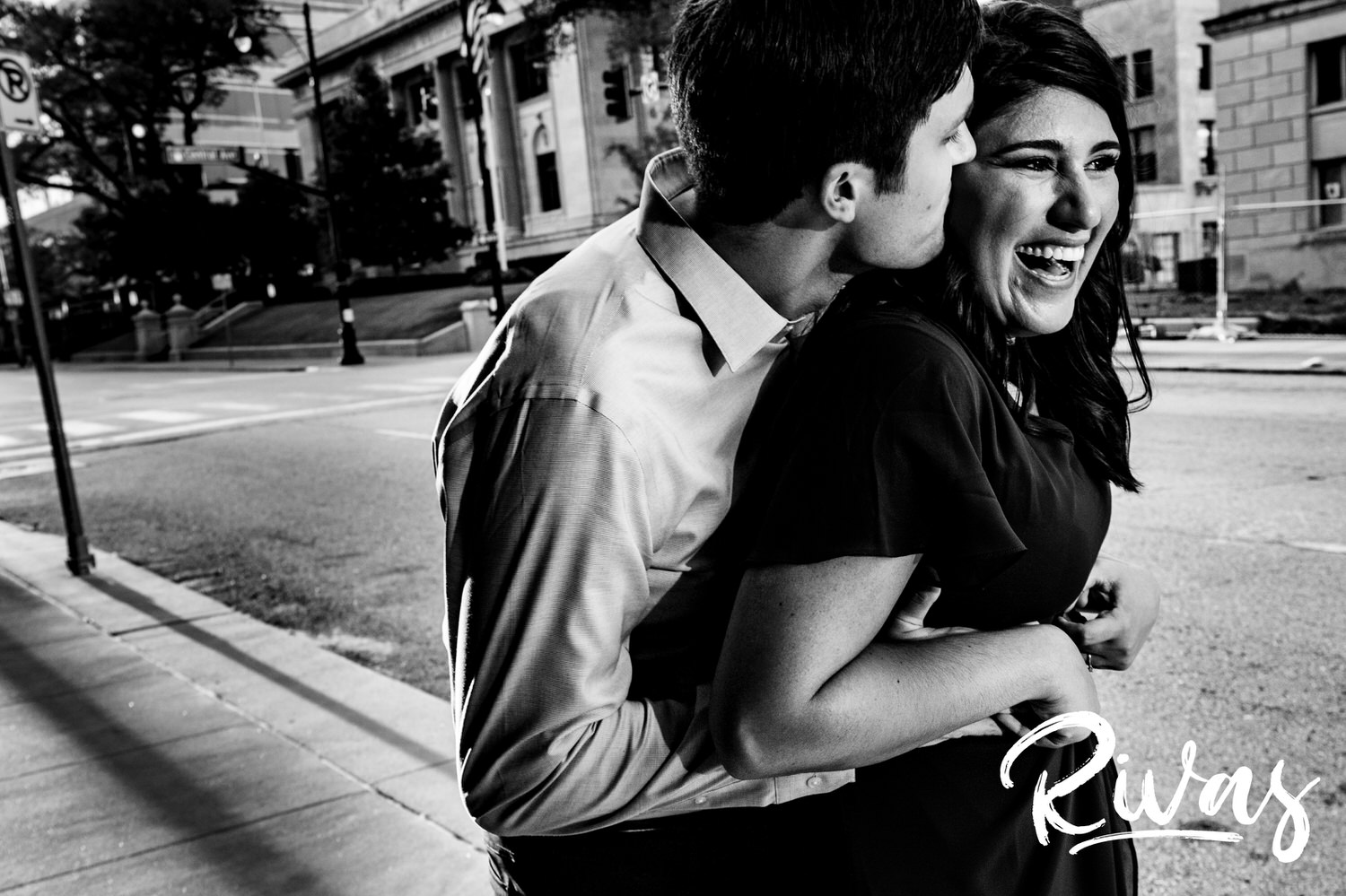 A candid black and white picture of an engaged couple sharing an embrace and whispering together during their downtown Kansas City fall engagement session. 