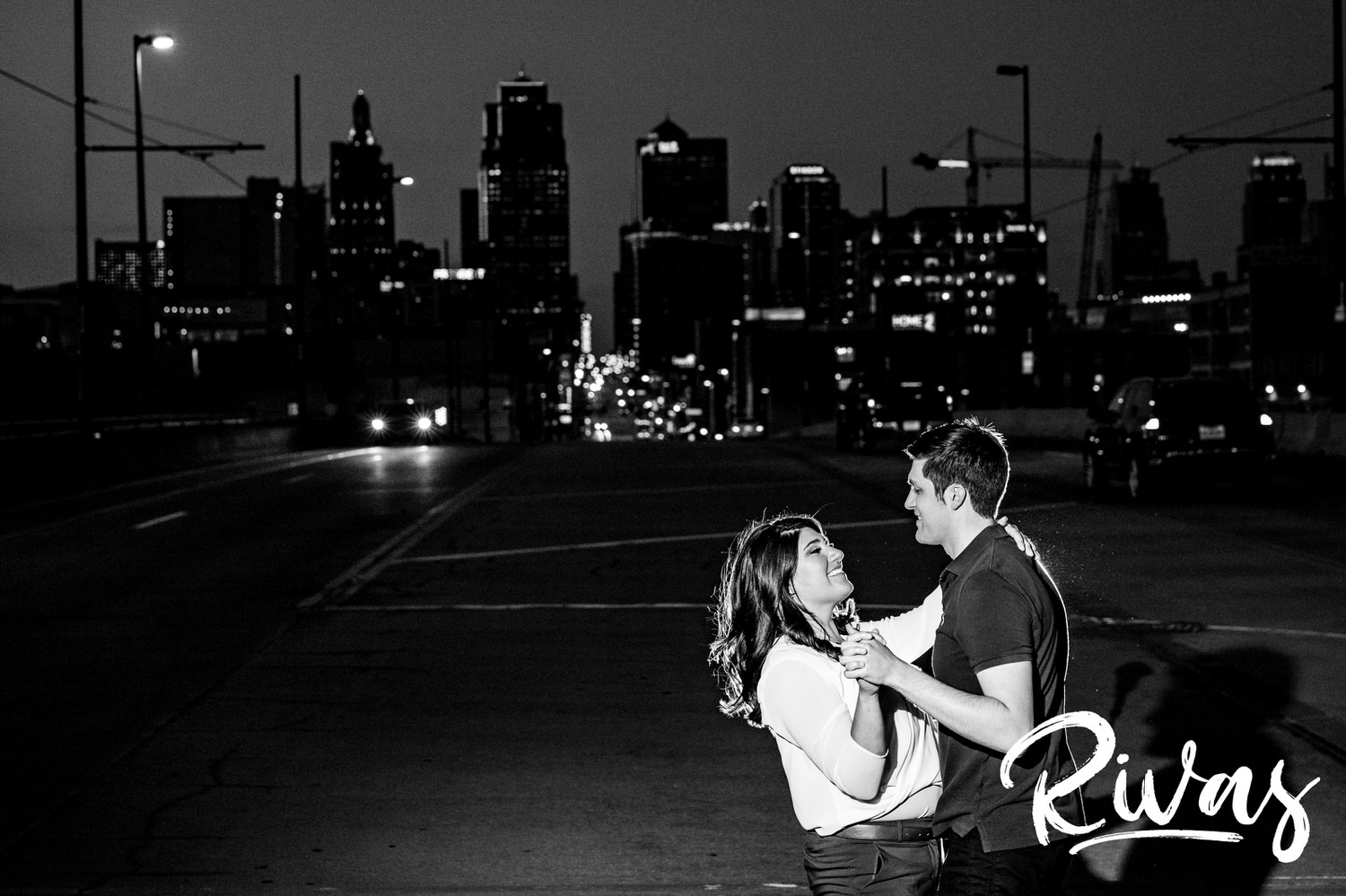A portrait of an engaged couple standing in the middle of Kansas City's Main Street, holding hands, just after sunset with the KC skyline visible behind them during their fall engagement session in Kansas City. 
