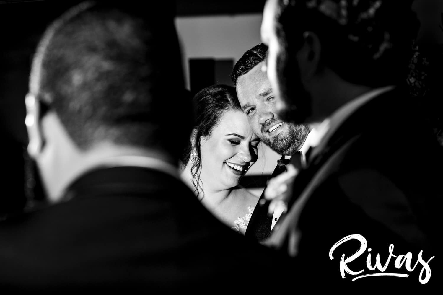 A candid picture taken through the shoulders of two groomsmen of a bride and groom leaning their heads together and laughing as they're toasted during their wedding reception at Lawrence Country Club in August. 