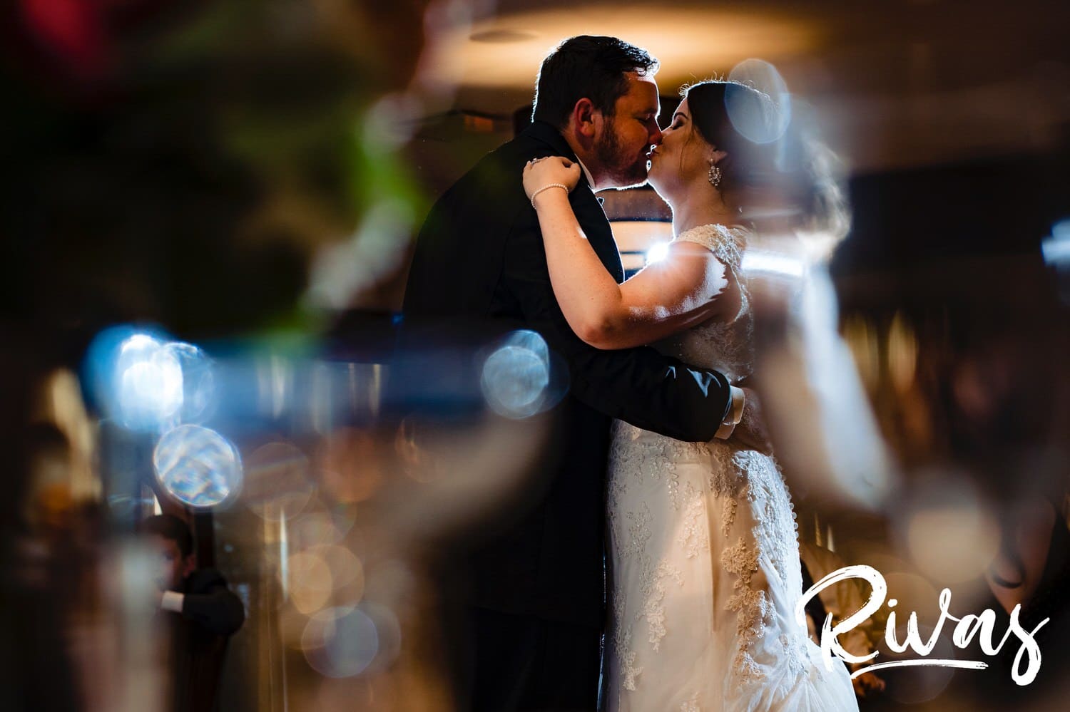 A candid picture taken through champagne glasses and floral arrangements on a table of a bride and groom sharing a kiss during their first dance at their summer wedding reception at Lawrence Country Club. 