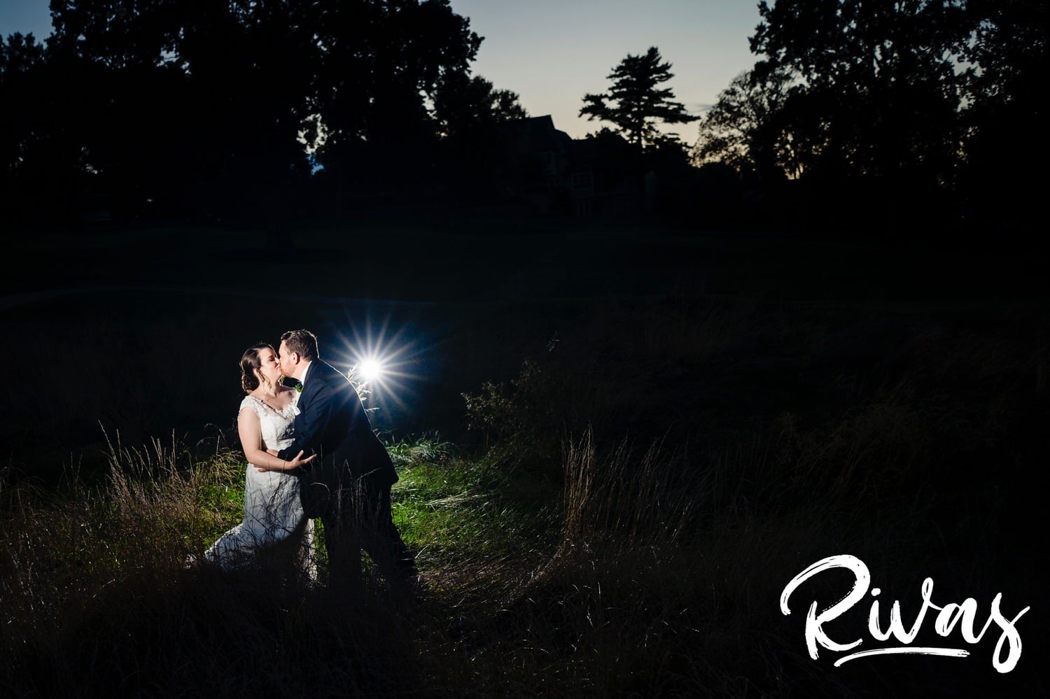 A candid picture of a groom kissing his bride and dipping her back just a little bit in super tall grass just after sunset on their summer wedding day at Lawrence Country Club. 