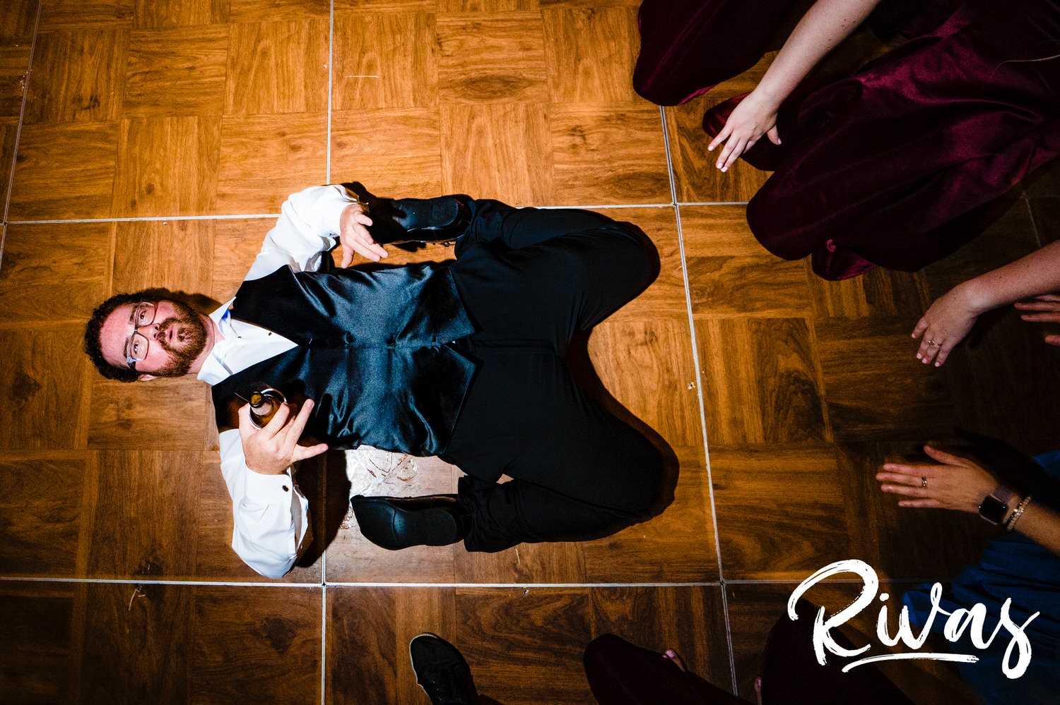 A candid picture taken from the top-down of a groomsman laying back on the dance floor, drink in hand as several people reach out to help him up during a summer wedding reception at Lawrence Country Club. 