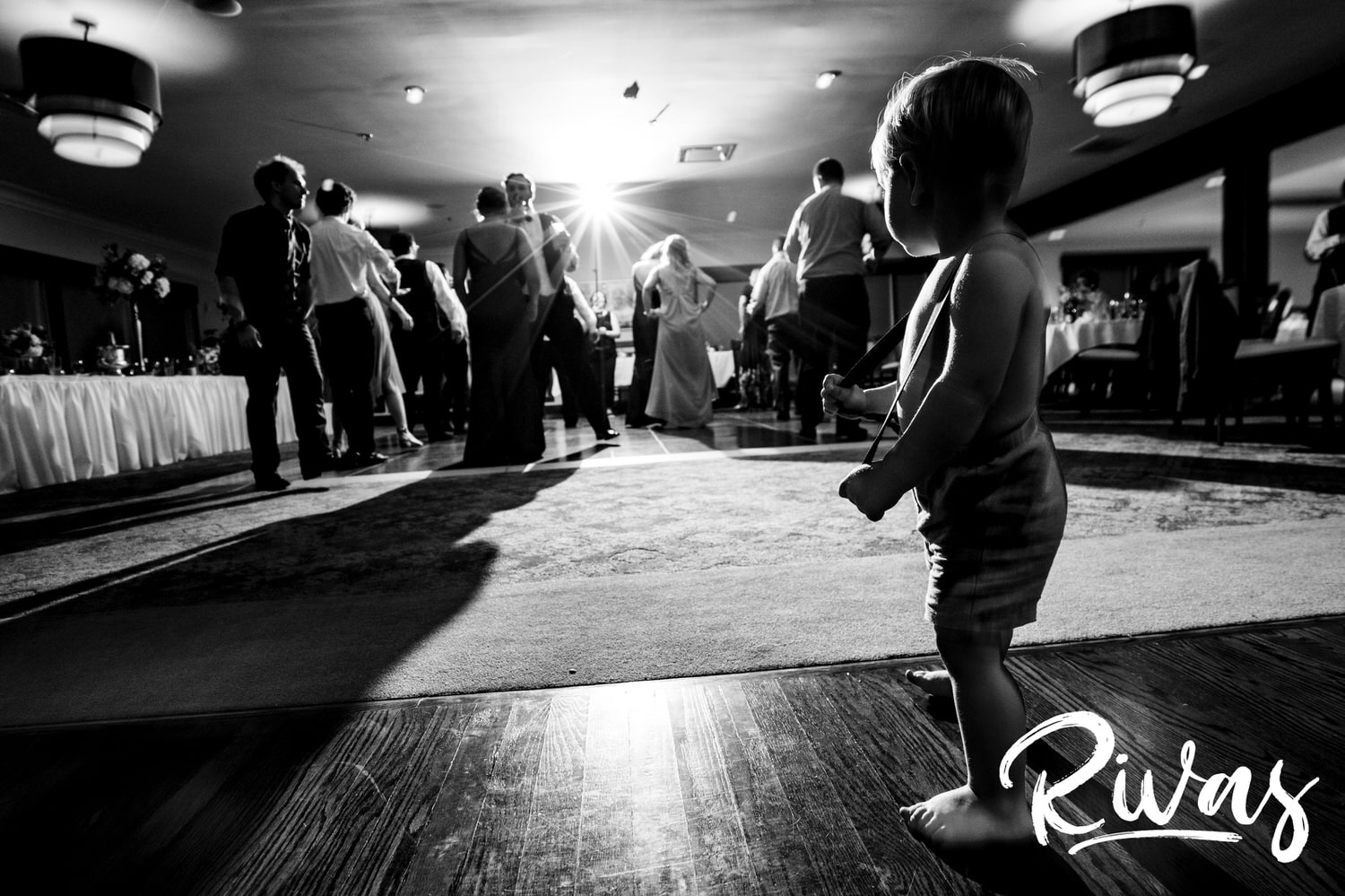A candid black and white picture of a baby in shorts and suspenders pulling his suspenders out as he walk in front of the full dance floor during a summer wedding reception at Lawrence Country Club. 