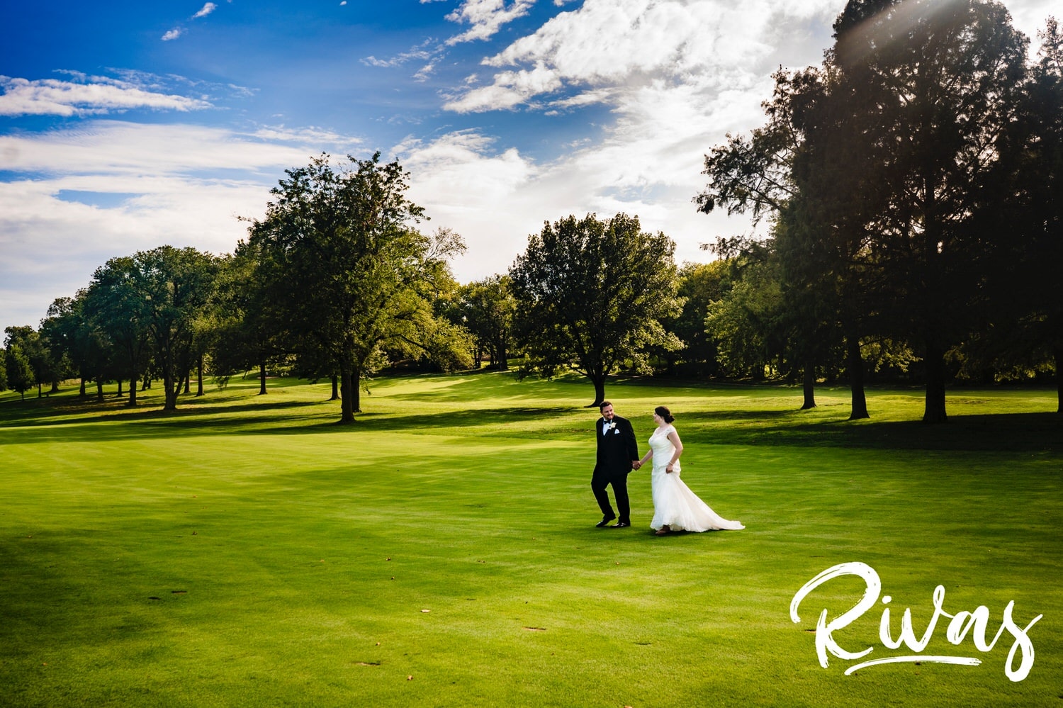A colorful, wide portrait of a bride and groom walking hand-in-hand across a putting green at Lawrence Country Club on their summer wedding day. 