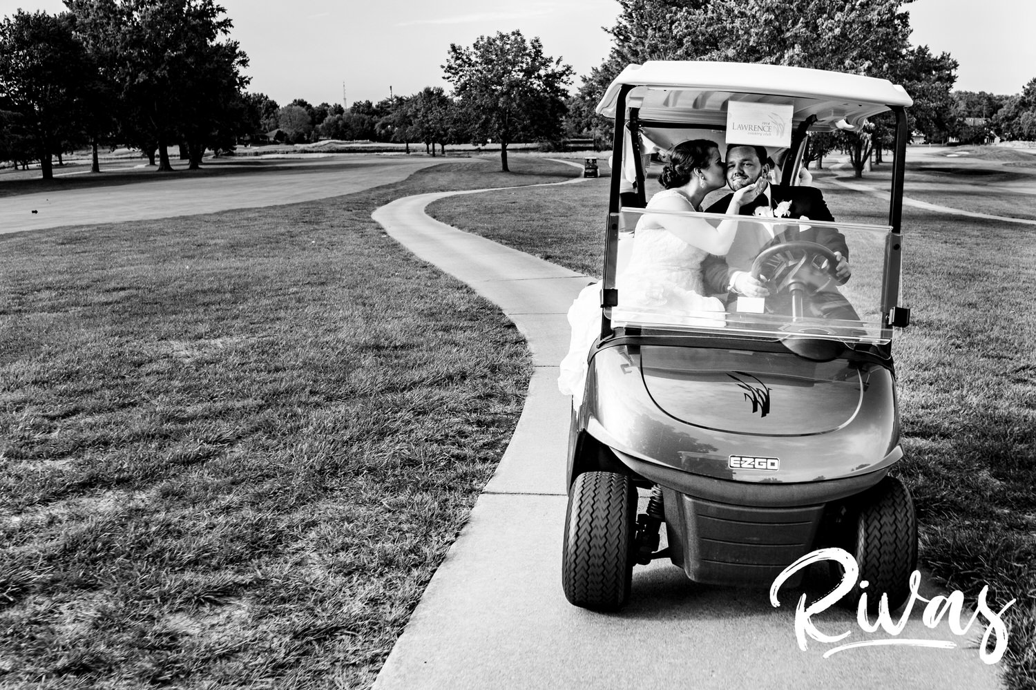 A candid black and white picture of a bride kissing her groom as he drives a golf cart across Lawrence Country Club on their hot summer wedding day. 