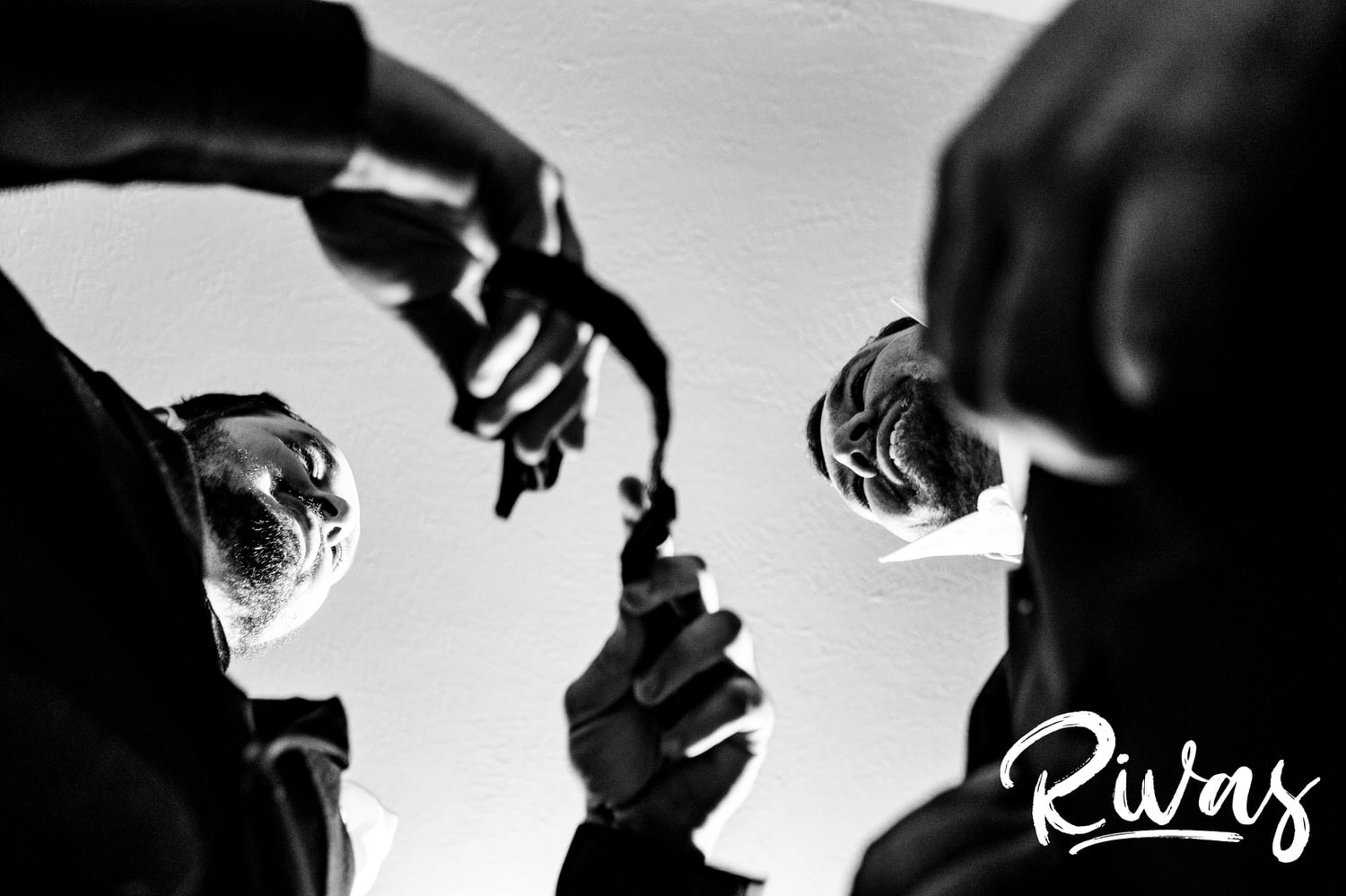 A candid black and white picture taken from below of a groomsman helping a groom put on his bowtie on the morning of his wedding in Lawrence, KS. 