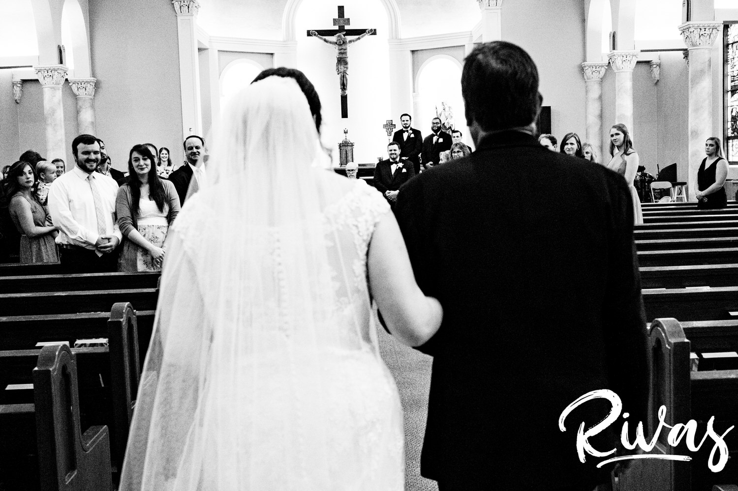 A candid black and white picture taken from over the bride's shoulder of her groom as her father escorts her down the aisle during her Lawrence, Kansas wedding. 