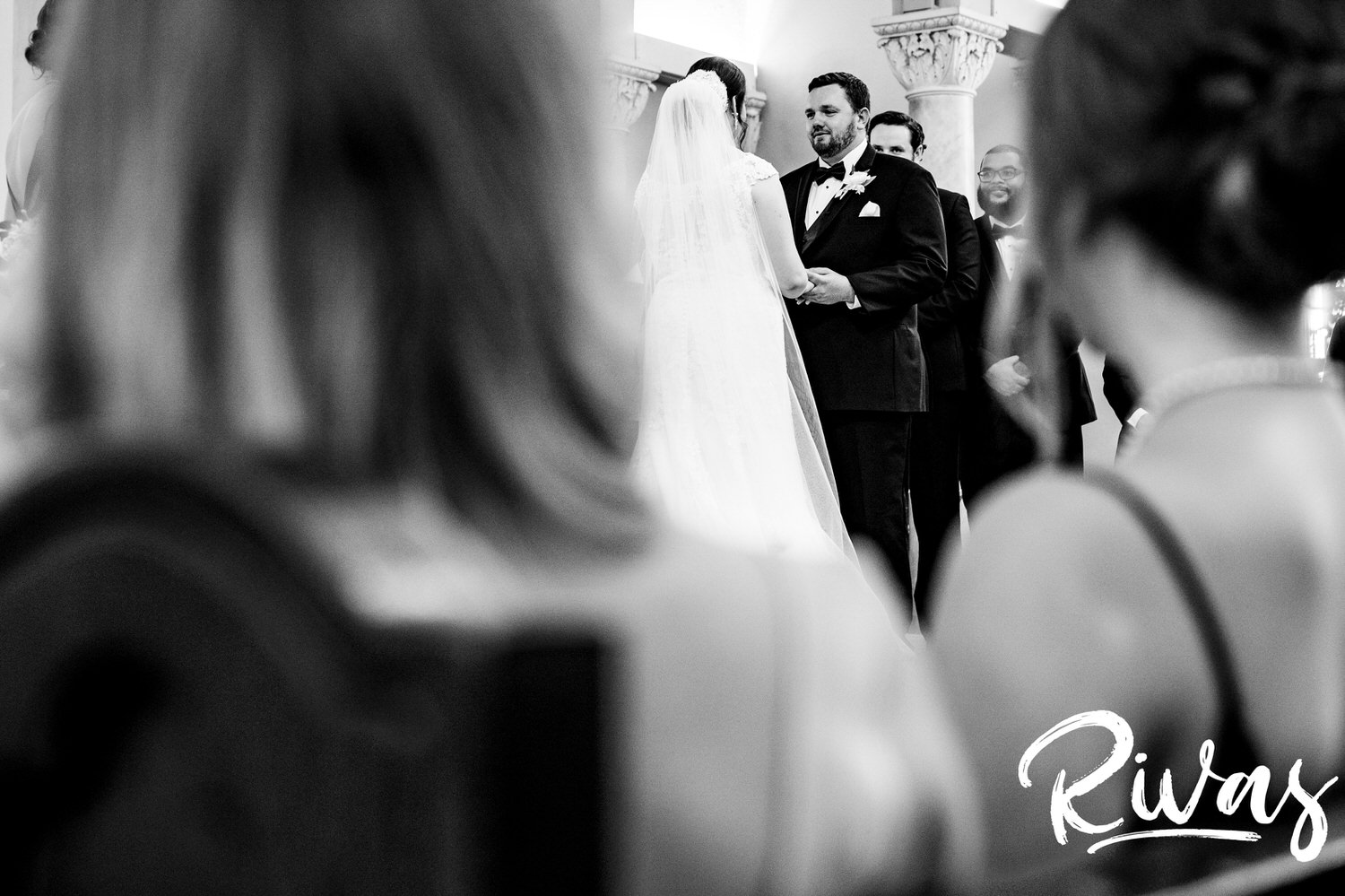 A candid picture taken through the shoulders of a group of bridesmaids of a bride and groom holding hands and exchanging vows during their wedding ceremony in Lawrence, Kansas. 
