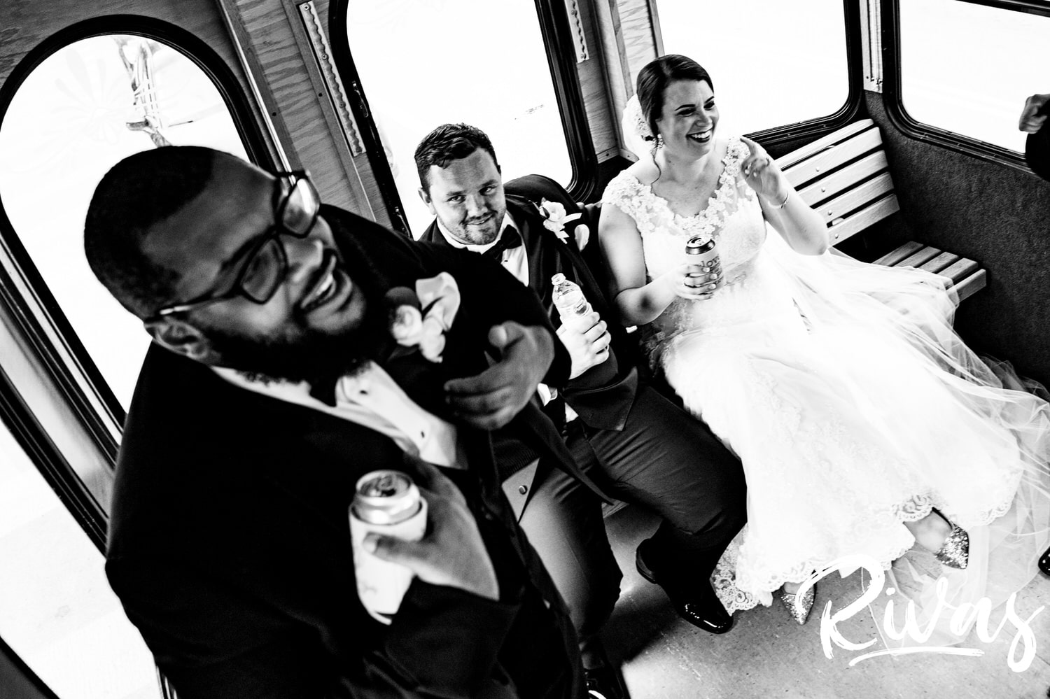 A candid black and white picture of a groomsman standing up  and singing as a bride and groom laugh and look on  while riding a trolley on their summer wedding day in Lawrence, Kansas. 