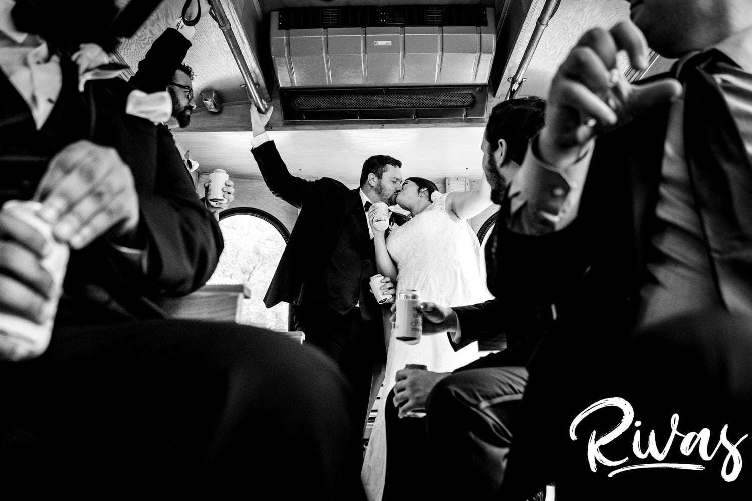 A candid black and white picture taken through the legs and arms of trolley riders of a bride and groom sharing a kiss on their summer wedding day in Lawrence, Kansas. 