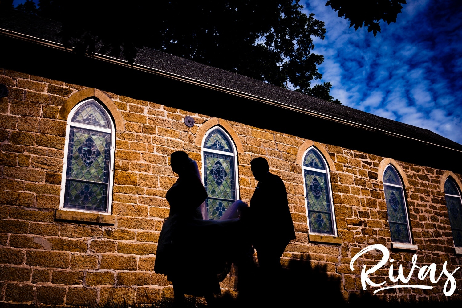 A colorful, silhouetted picture of a groom carrying his bride's train across a yard in front of an old church on their summer wedding day in Lawrence, Kansas. 