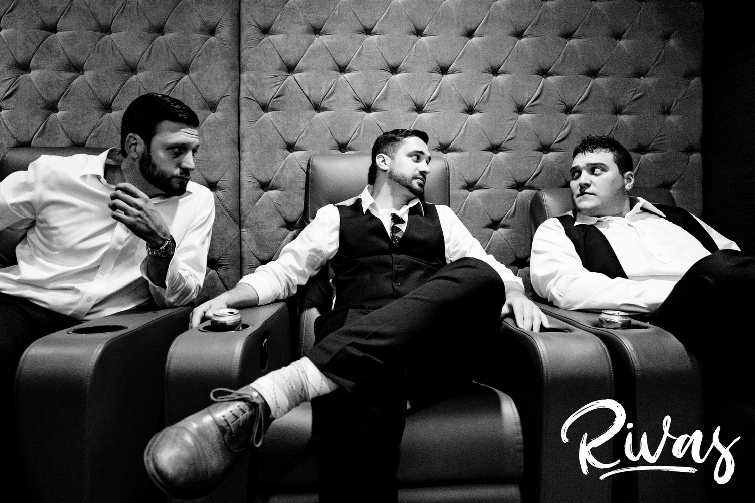 A candid black and white picture of a groom and two groomsmen, sitting in chairs, reclining and making funny faces at each other. 