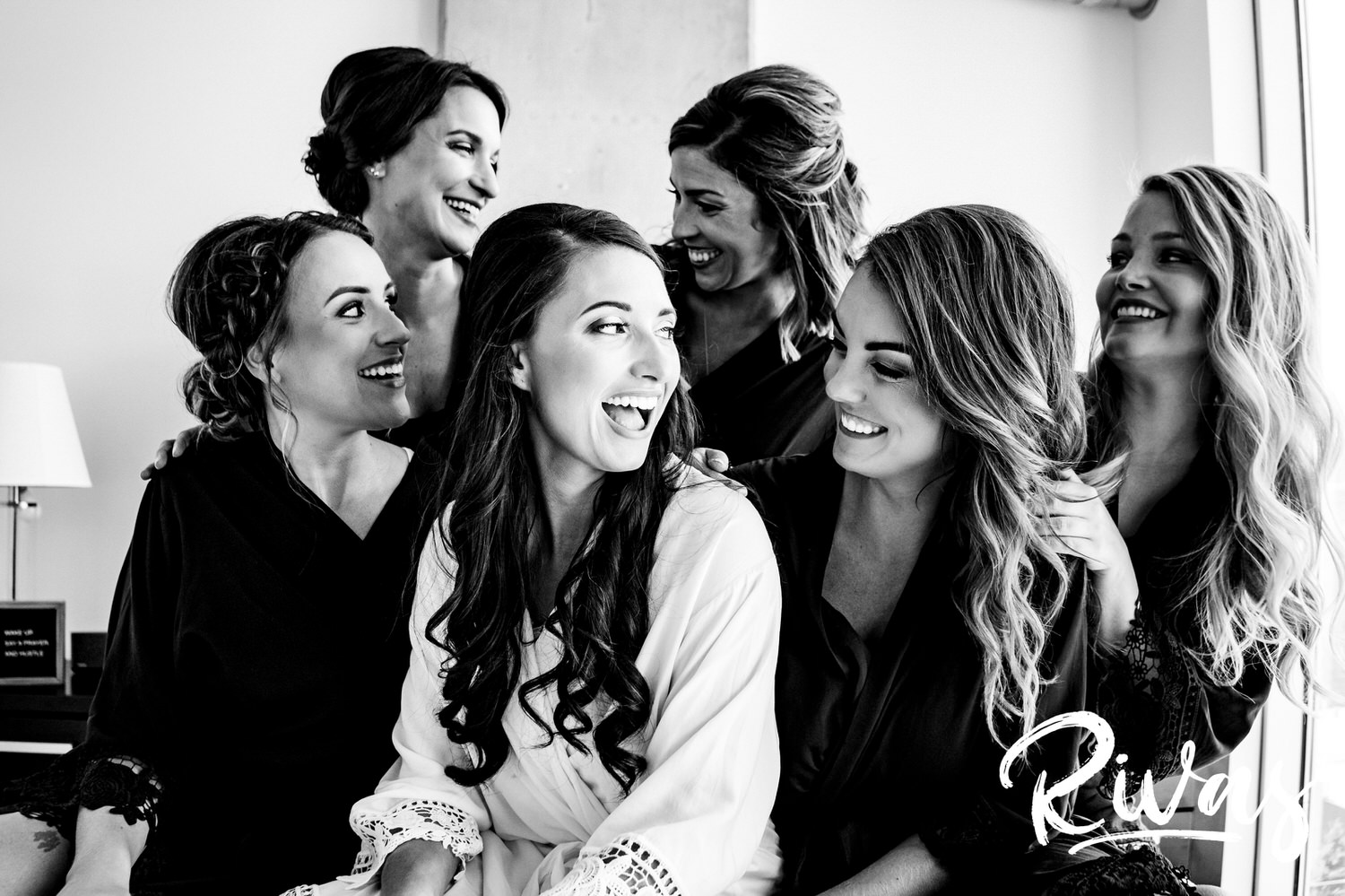 A candid black and white picture of a bride and her bridesmaids sitting on a bed, laughing together  on the morning of a wedding in downtown Kansas City. 