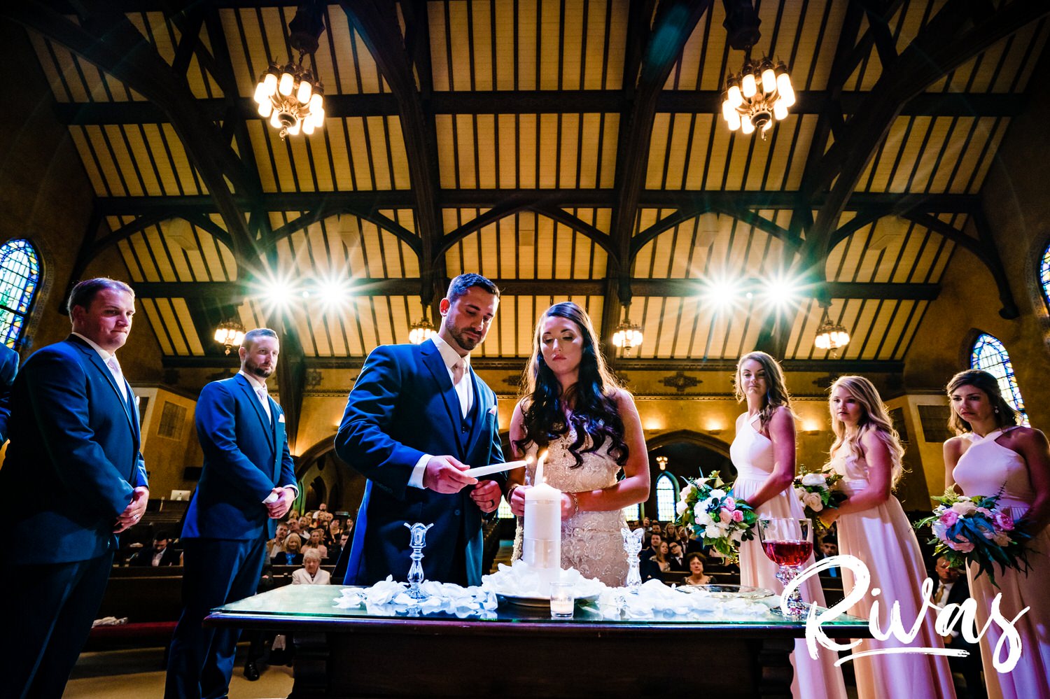 A colorful, candid picture taken from behind the altar of a bride and groom leaning together lighting their unity candle during their wedding ceremony. 