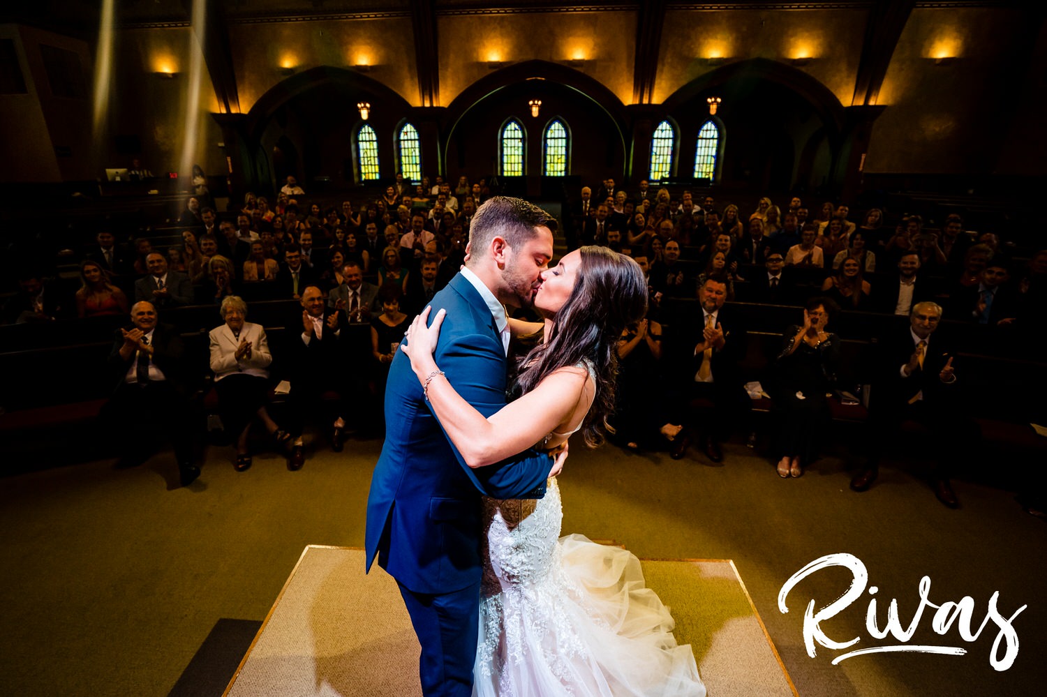 A candid picture taken from behind the bride and groom, looking out at the audience, as they share their first kiss at the end of their wedding ceremony in Kansas City. 