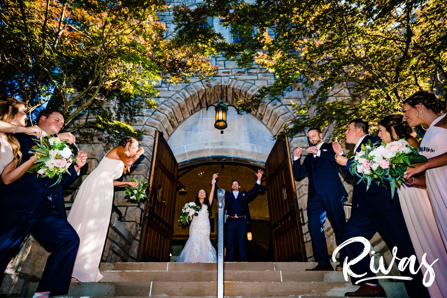 A candid picture, taken from the front of the church of a bride and groom holding hands in celebration as they walk down the stairs to a flurry of bubbles after their wedding ceremony. 