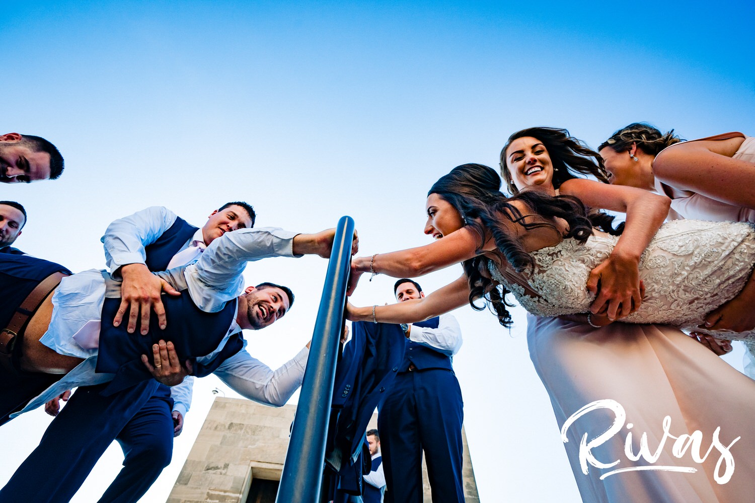 A candid, colorful picture of a bride and groom, holding on to a rail, as their bridal party pulls them apart on their wedding day. 