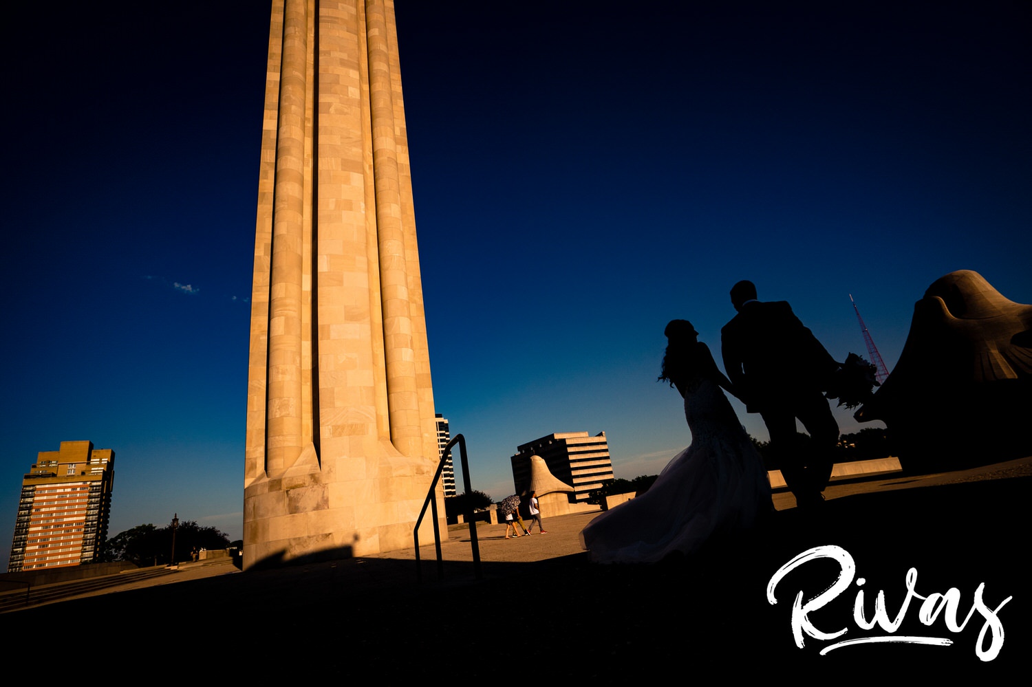 A dramatic, colorful picture of a bride and groom, mainly in the shadows, of a bride and groom holding hands and walking across the base of Liberty Memorial on their Kansas City wedding day. 