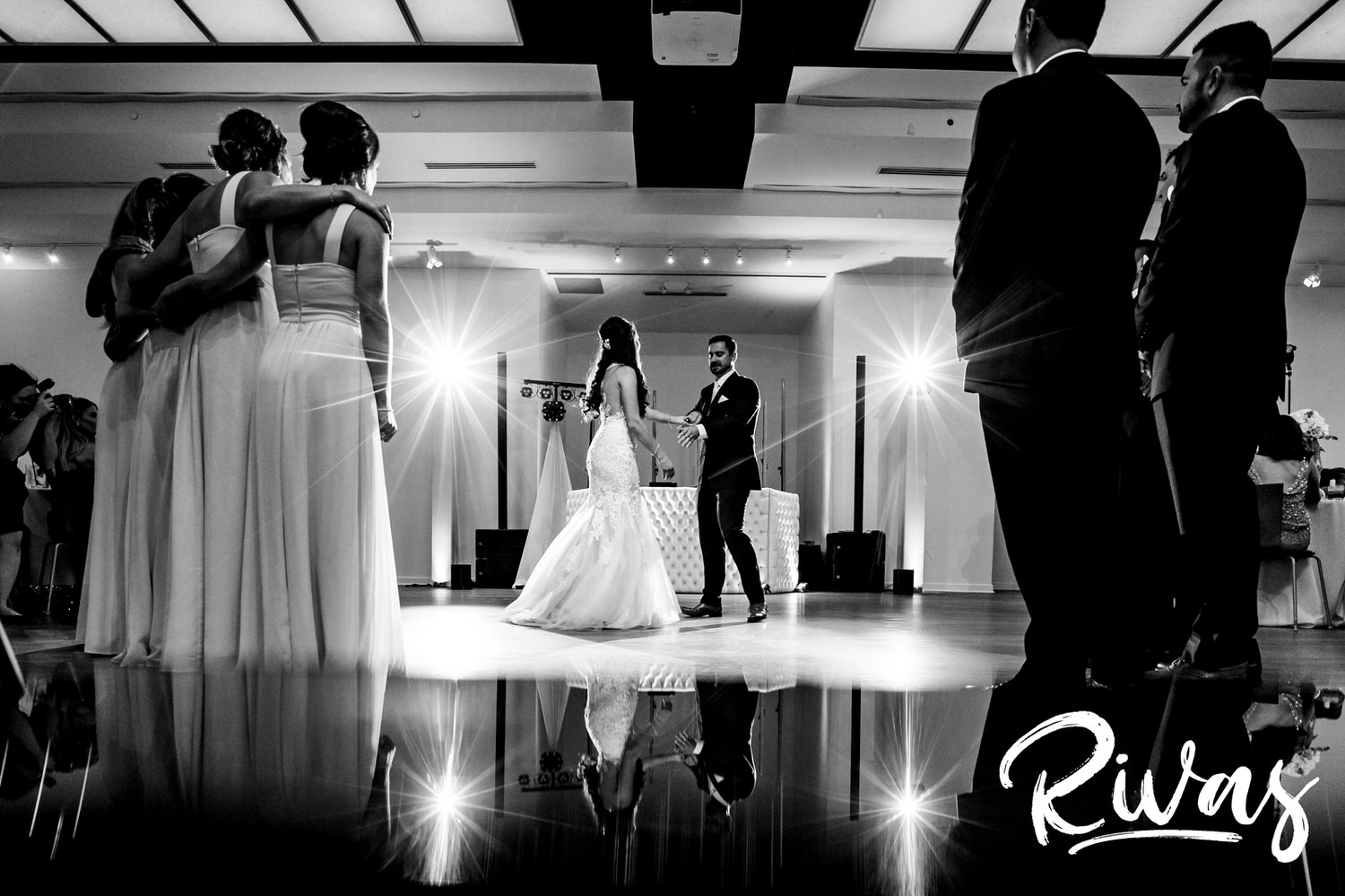 A candid black and white picture of a bride and groom sharing their first dance, as their wedding party stands watching during a wedding reception at The Gallery in downtown Kansas City. 