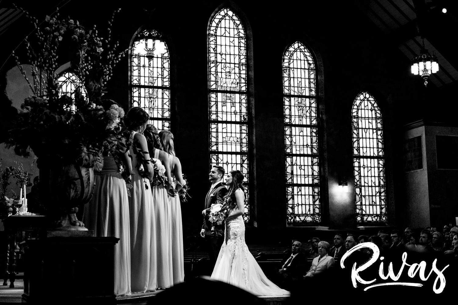 A candid, black and white, wide picture, taken from the side of the altar of a bride and groom standing on stairs looking up at their officiate, in front of a row of stained glass windows during their wedding ceremony in Kansas City. 
